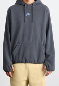 Grå fleecetröja med huva och känguruficka, snörning vid huvan och blå Nike-logotyp på bröstet. Har ribbstickade muddar och fåll.