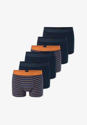 Uomo senza maglietta con capelli corti, braccia incrociate, che indossa boxer blu navy con strisce orizzontali arancioni e una fascia in vita arancione.