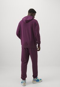 Felpa con cappuccio viola e pantaloni coordinati, realizzati in tessuto morbido. Presenta una vestibilità comoda, polsini a coste e sneakers bianche con accenti blu e rosa.