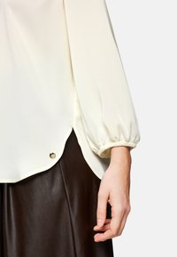 Cremfarbener Blouson-Oberteil mit weit ausgestellten Ärmeln, abgerundetem Saum und goldenen Hardwaredetails an der Seite, kombiniert mit einem dunkelbraunen Lederrock.