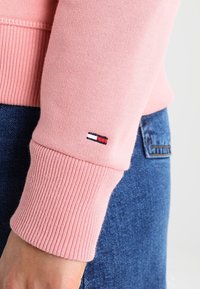 Sudadera rosa con puños acanalados, que presenta un pequeño logo bordado en azul, rojo y blanco en la manga. Fondo de mezclilla visible.