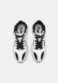 Zapatillas deportivas en blanco y negro con detalles de malla, suelas de goma y diseño de puntera redonda. Presentan cordones negros y un acabado beige neutro.