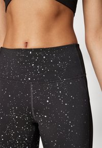 Zbliżenie na środek ciała osoby noszącej czarne legginsy z odbijającym srebrnym, plamkowanym wzorem oraz dopasowaną czarną bluzkę sportową.