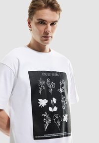Camiseta de algodón blanca con un estampado gráfico de flores en arte lineal negro. Diseño de manga corta y cuello redondo. Textura plana sin adornos.