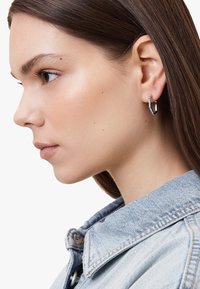 Orecchino a cerchio in argento con design geometrico, decorato con cristalli trasparenti lungo un bordo. Chi lo indossa ha i capelli castani lisci e indossa una giacca di jeans.