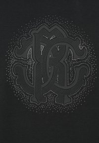 Czarna koszulka z wyraźnym, teksturowanym logo z ozdobnymi ćwiekami o różnych rozmiarach wokół krawędzi, tworząca migoczący efekt.