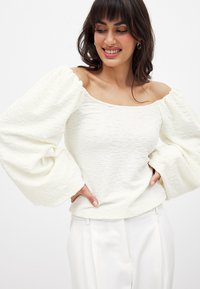 Top bianco con texture, scollatura off-shoulder e maniche a sbuffo voluminose. Il tessuto appare leggero e morbido, abbinato a pantaloni bianchi su misura.
