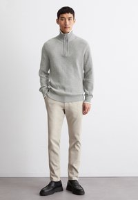 Grey knitted sweater with a zip-up collar, paired with beige trousers and black loafers, standing against a light background.