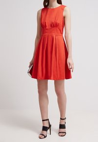 Donna che indossa un vestito rosso senza maniche, lungo fino al ginocchio, con una gonna plissettata, mentre tiene in mano una pochette argentata e sandali neri con tacco alto e cinturino alla caviglia.