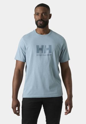 Hombre con barba vistiendo una camiseta azul claro de Helly Hansen y pantalones negros, de pie frente a un fondo claro y uniforme.