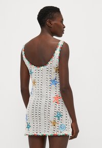 Vestido de ganchillo blanco con detalles de aplicaciones en forma de estrellas de mar y espirales de colores. Presenta un escote en la espalda y un borde decorativo en tonos pastel.