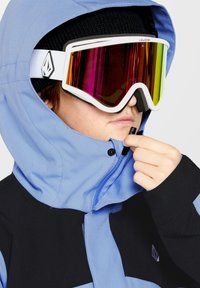 Skibril met een wit frame en kleurrijke reflecterende lens. De drager draagt een blauw en zwart capuchonjack en past de rits aan.