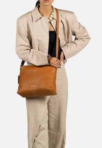Sac en cuir brun à bandoulière de forme rectangulaire, présentant une surface texturée et un logo en détail. La sangle est réglable pour plus de commodité.