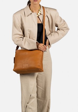 Sac en cuir brun à bandoulière de forme rectangulaire, présentant une surface texturée et un logo en détail. La sangle est réglable pour plus de commodité.