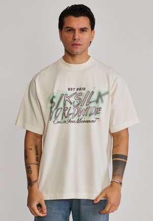 Homme aux cheveux foncés portant un T-shirt blanc ample avec un graphisme et un jean bleu, debout avec une expression neutre sur un fond uni.
