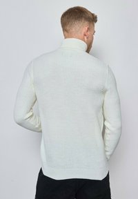 Pull en maille blanche avec un col haut, présentant une texture côtelée et des manches cintrées. Le dos est uni, mettant en valeur le motif tricoté.