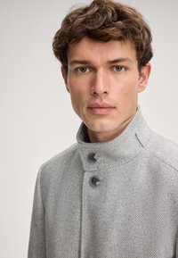 Jonge man met bruin krullend haar die een lichtgrijze hogenekjas draagt met twee zichtbare knopen, recht vooruit kijkend tegen een effen achtergrond.