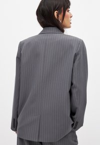 Pelēkas svītrains žakete ar pielāgotu piegriezumu, vienrindas dizainu, izgrieztiem apkakles galiem un pogātām piedurknēm, demonstrējot gludu tekstūru.