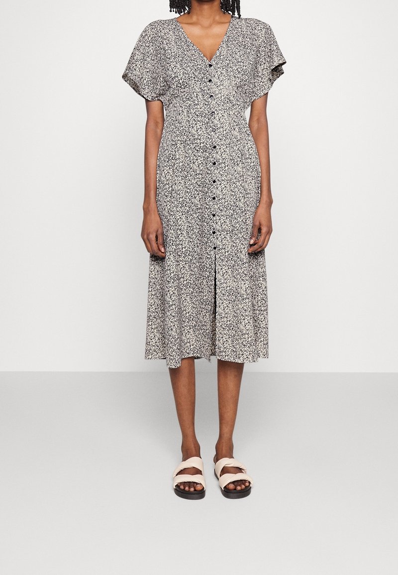 Černobílé vzorované šaty po kolena s výstřihem do V, krátkými volánovými rukávy a knoflíky vpředu, doplněné světlými sandály.