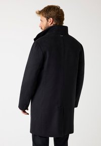 Lungo cappotto nero realizzato in tessuto morbido, con colletto alto, vestibilità dritta e tasche laterali. Sul retro presenta un dettaglio di cucitura centrale.