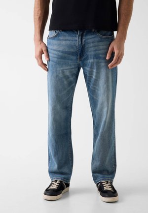 Mężczyzna w wyblakłych, prostych niebieskich dżinsach i czarnych trampkach z białymi sznurowadłami, stojący z rękami luźno opuszczonymi wzdłuż ciała.