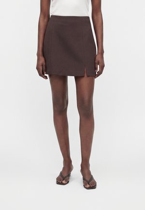 Femme portant une mini-jupe marron foncé avec une fente à l'avant, un haut blanc et des sandales à talons ouvertes noires, debout sur un fond uni.