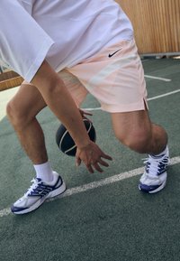 Nike træningsshorts i lys pink mesh med hvide detaljer og et sort logo. Hvide træningssko med blå og lilla detaljer, holder en basketball.