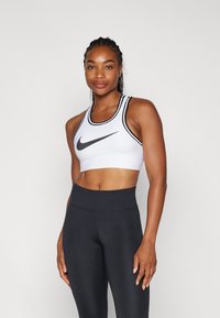 Hvit sports-bh med svarte detaljer og teksturert stoff, med en fremtredende Nike-logo. Kombinert med sorte high-waisted leggings.
