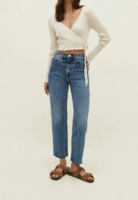 Nainen, jolla on päällä vaalean kermanvärinen kietaisupaita, korkeavyötäröiset siniset farkut raidoilla, ja ruskeat kaksinauha-sandaalit, seisoo yksinkertaista taustaa vasten.