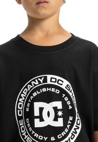 Černé bavlněné tričko s kulatým bílým logem obsahujícím "DC Shoe Company," založenou v roce 1994, a textem "Destroy & Create."