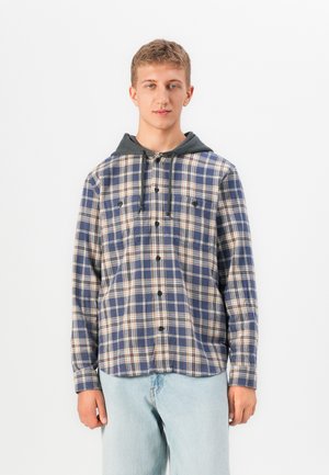 Jonge man in een blauw en beige geruit button-up shirt met een grijze capuchon, gecombineerd met lichtblauwe jeans, staande tegen een witte achtergrond.