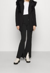 Kobieta ubrana w czarną pikowaną kurtkę z futrzanym kapturem, białą bluzkę, czarne spodnie w stylu flare z rozcięciem oraz białe botki na kostkę, stojąca na szarym podłożu.