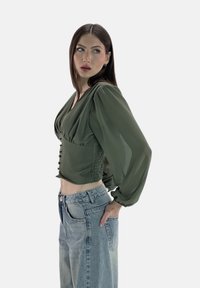 Zielona oliwkowa bluzka z bufkami, dekoltem w serek i detalami w postaci guzików, połączona z jasnoniebieskimi dżinsami o wysokim stanie. Regulowane dopasowanie.