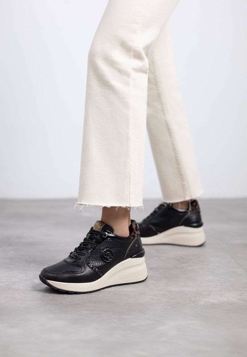 Zapatillas sintéticas negras con detalles de malla, suela en beige tipo cuña y detalles de leopardo en el talón, combinadas con jeans color crema.