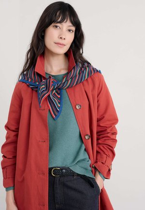 Rode jas met knopen, groene gebreide trui met textuur, en een gestreepte sjaal in blauw, rood, groen en geel. Zwarte gevlochten riem als accent op donkerblauwe jeans.