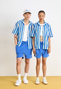 Gestreepte shirts met korte mouwen in blauw en beige, gecombineerd met blauwe shorts. Katoenen materiaal met zichtbare logo patches. Witte schoenen.