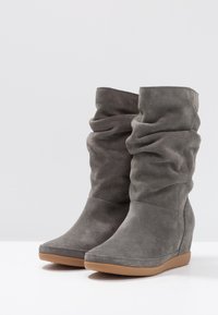 Stivali con zeppa in suede grigio dal design sfilacciato. Presentano una punta rotonda e una superficie texturizzata. La suola in gomma marrone crea un contrasto.