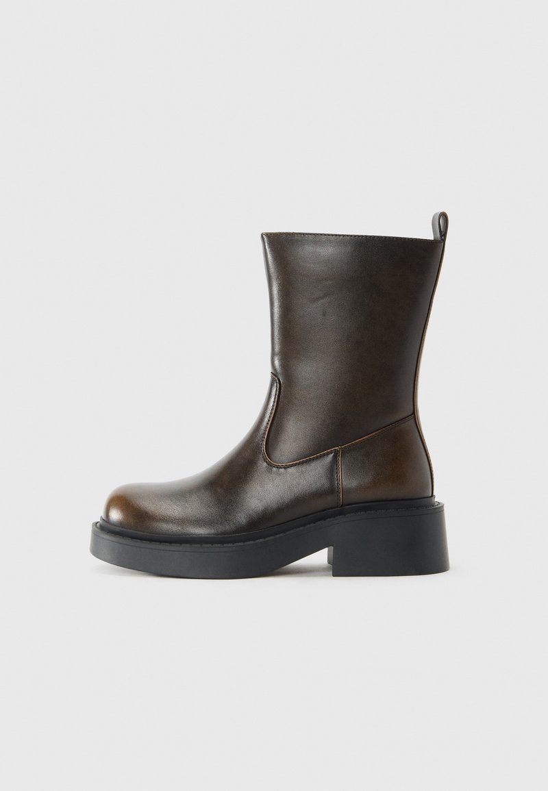 Botte en cuir marron foncé mi-mollet avec bout arrondi, semelle épaisse noire, petit talon et languette à l'arrière sur fond clair uni.