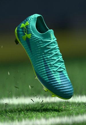 Cyaan en limoen groene voetbalschoen met veters, in de lucht boven het gras op een gemarkeerd voetbalveld, met grasresten die in de lucht zweven.