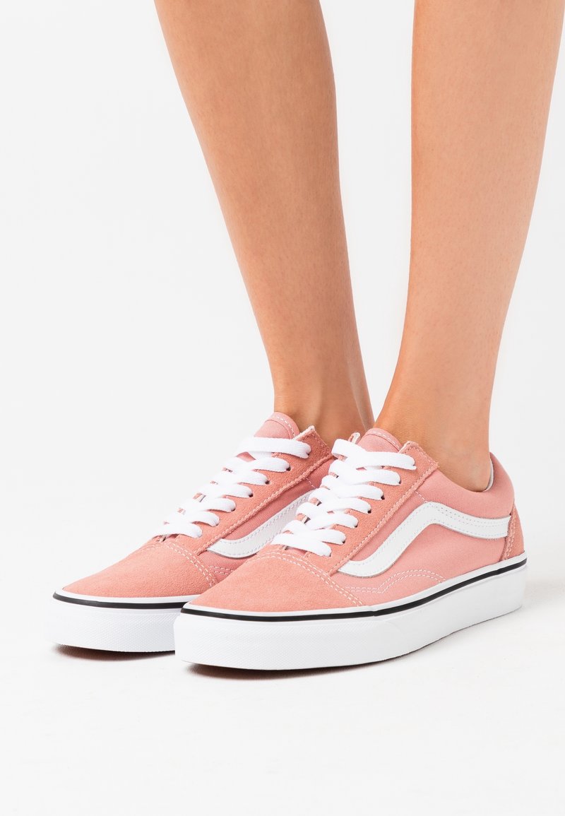 Pieds portant des baskets basses en daim rose avec des lacets blancs et une bande latérale blanche sur fond blanc.