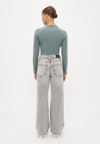 Top de manga larga de color verde claro combinado con unos vaqueros de pierna ancha de talle alto en gris claro. La parte posterior presenta un escote curvado y dos bolsillos traseros.