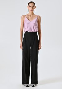 Camicetta di raso rosa chiaro con spalline sottili abbinata a pantaloni neri a vita alta. I tacchi neri presentano dettagli a fiocco argentati. Sfondo semplice.