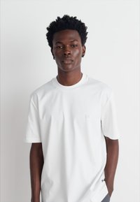 Tricou alb cu mânecă scurtă, dintr-un material moale, având un logo subtil pe piept și o croială largă. Textură netedă și design clasic.