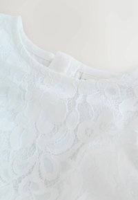 Valge pitsist topp lillemustriga, millel on sujuv kaelus ja kangast sildike. Tekstuur on õrn ja keeruline.