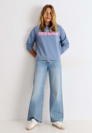Mujer con pelo rubio que lleva una sudadera azul con el texto "BROWNIE", vaqueros anchos de color azul claro y zapatillas blancas, de pie junto a una pared blanca.