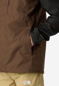 Main dans la poche d'une veste marron avec un poignet noir portant l'étiquette "DRYVENT", portant un pantalon beige avec le logo blanc "The North Face" sur la cuisse droite.