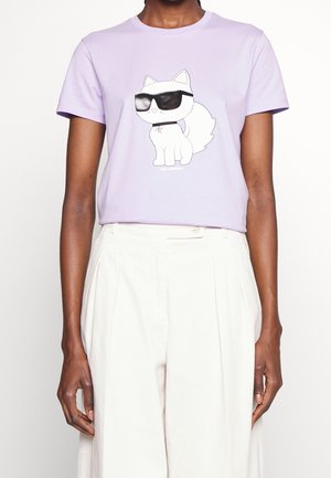 Persona que lleva una camiseta lavanda con un perro de dibujos animados blanco con gafas de sol negras estampado, combinada con pantalones blancos de cintura alta.