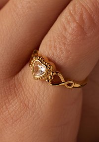 Goldring mit einem herzförmigen klaren Edelstein, aufwendigen Perlendetails und einem gewundenen Banddesign, vor einem strukturierten Hauthintergrund.
