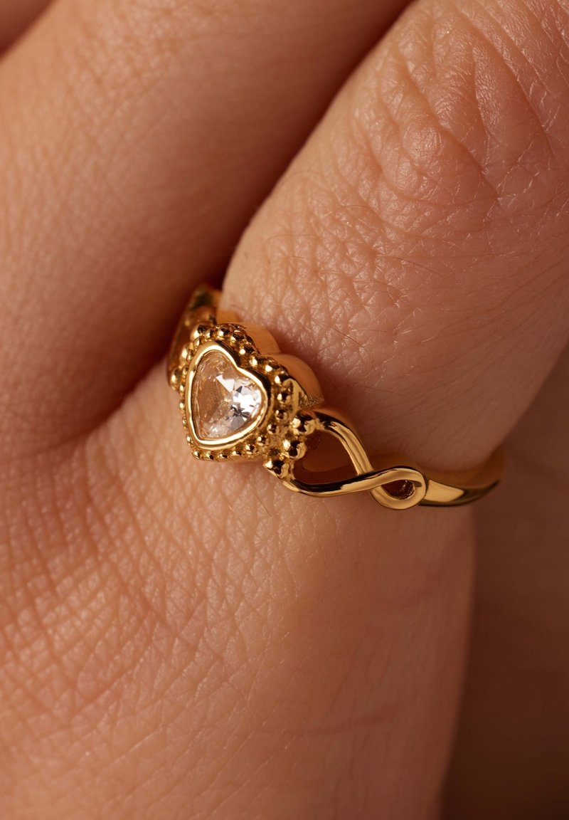 Goldring mit einem herzförmigen klaren Edelstein, aufwendigen Perlendetails und einem gewundenen Banddesign, vor einem strukturierten Hauthintergrund.