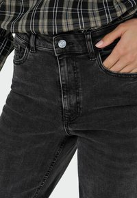 Črne kavbojke z visokim pasom, ki imajo klasično zapiranje z gumbom in stranske žepke, ter prikazujejo gladko, rahlo zbledelo teksturo.